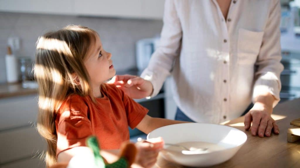 A young girl looks up at an adult in a kitchen while eating.