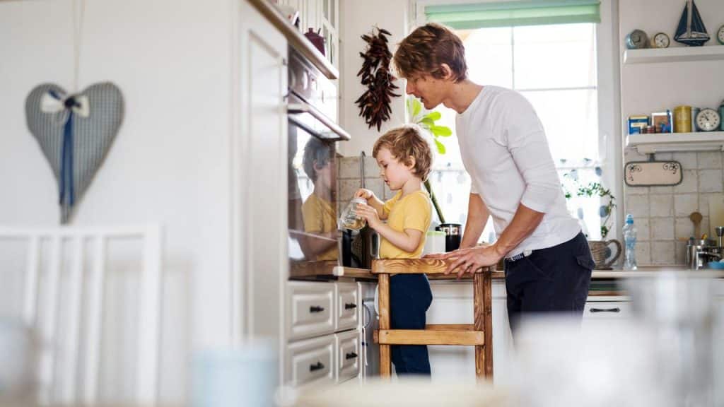 A young boy stands on a stool in a kitchen while an adult helps him.