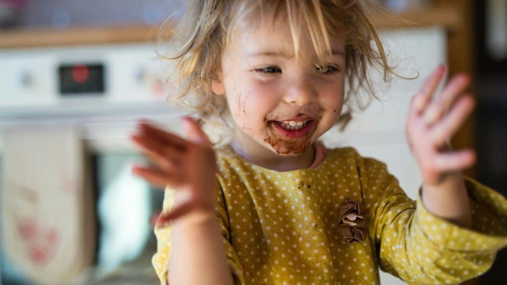 A young girl with a messy face smiles while playing in a kitchen.