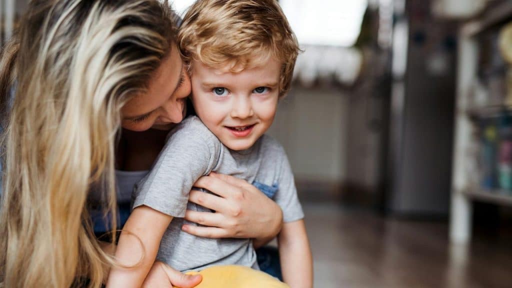 A young boy smiles as his mother hugs him gently.