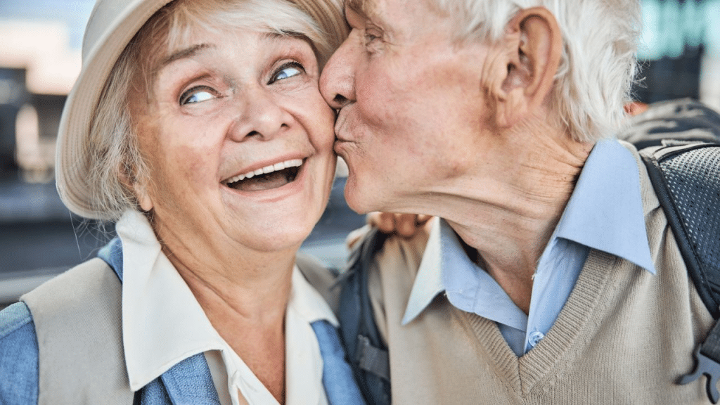 A smiling older woman in a hat is kissed on the cheek by an older man.