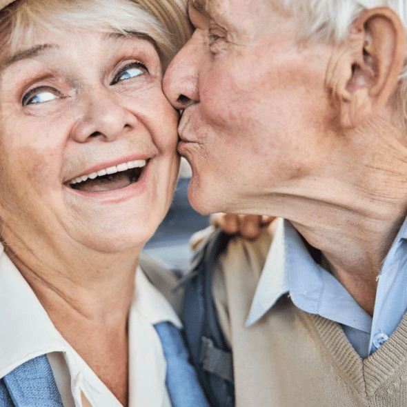 A smiling older woman in a hat is kissed on the cheek by an older man.