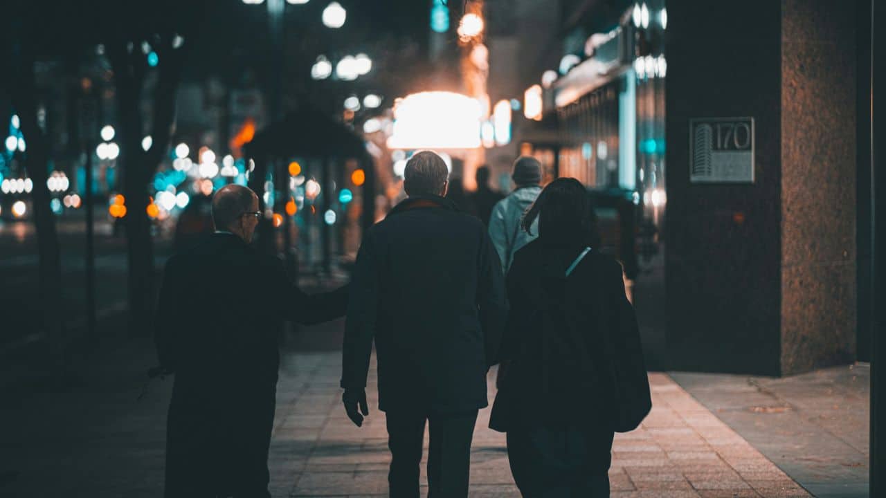 A group of three people walks down a bustling city sidewalk at night with blurred lights in the background.