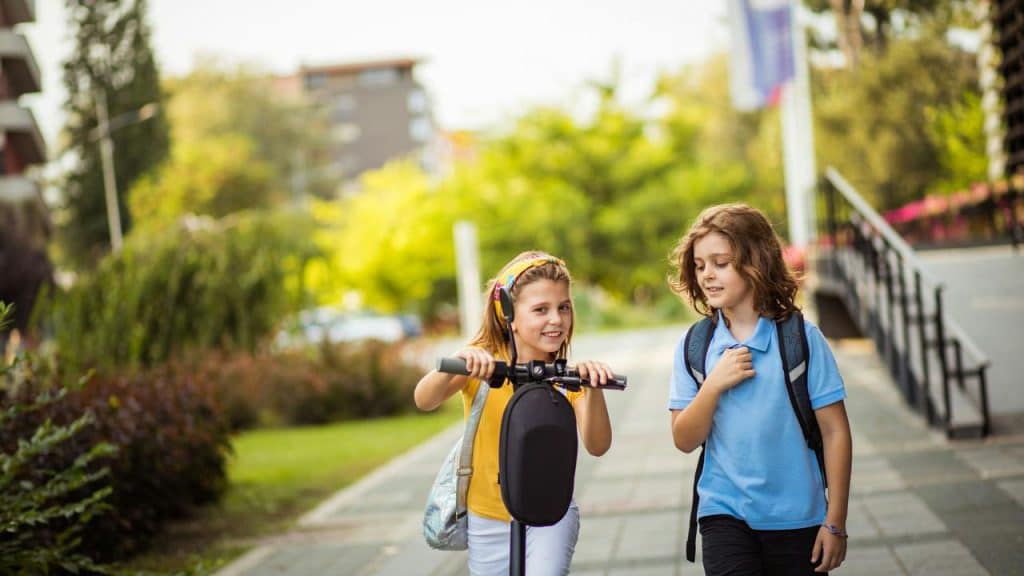 A photo of two kids outside, one walking and one riding a scooter.