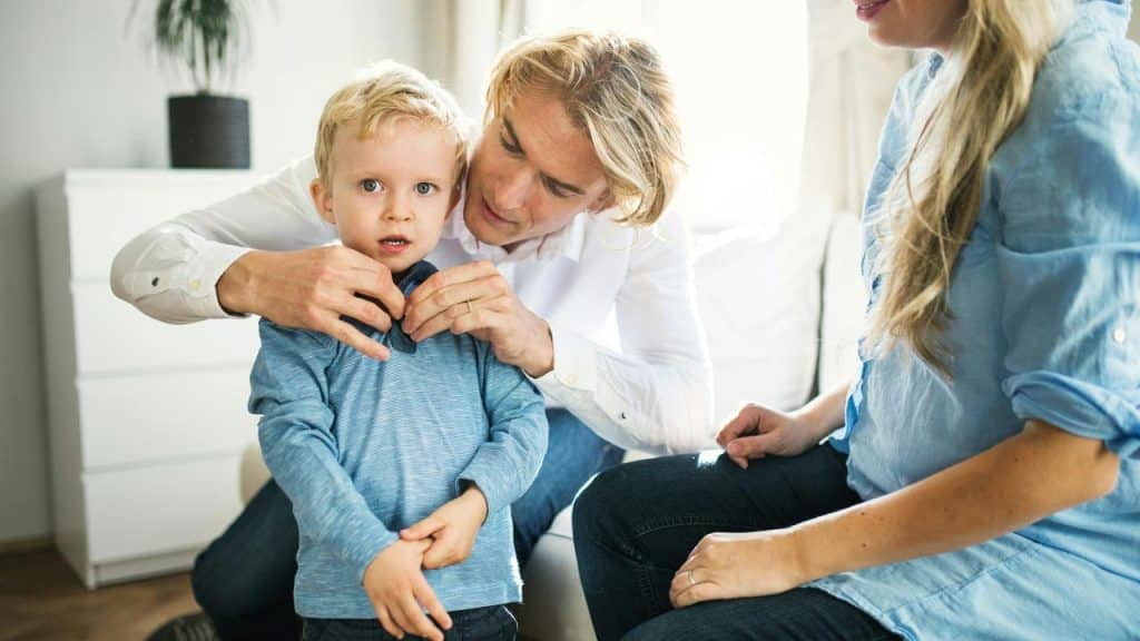 A father fixing his sonโs shirt as his mother watches from the side.