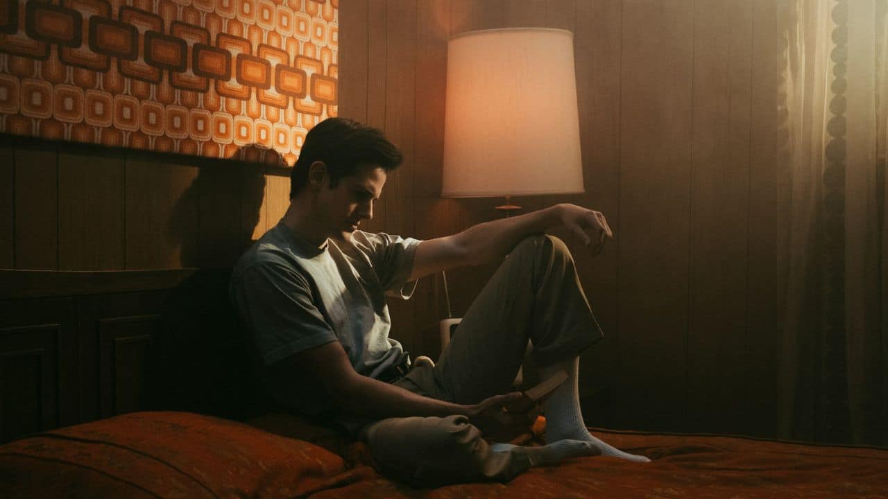 A young man in a retro-style room sits on a bed, looking down at his phone.