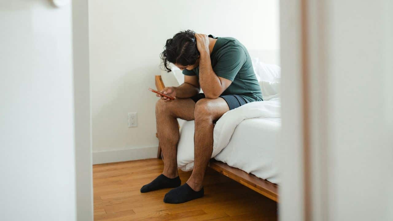 A distraught man sits on the edge of a bed, looking down at his phone with a hand on his head.