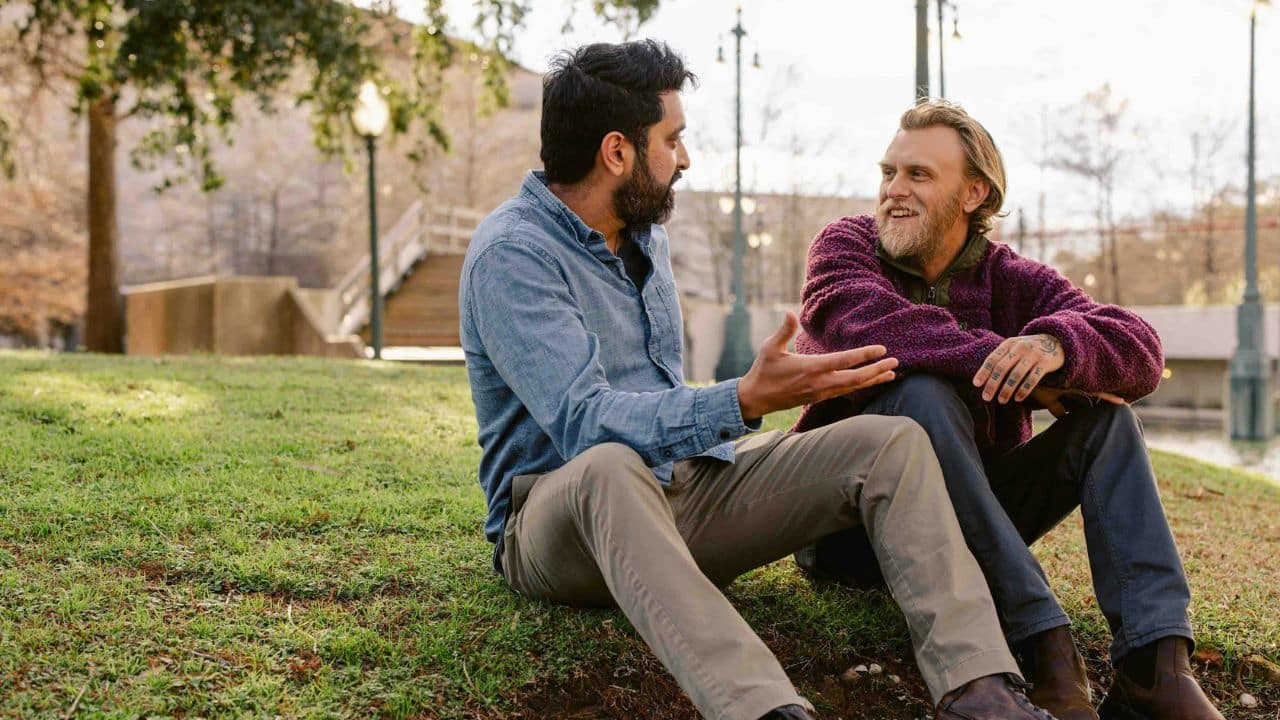 Two men sit on a grassy hill, engaged in a conversation in a park.