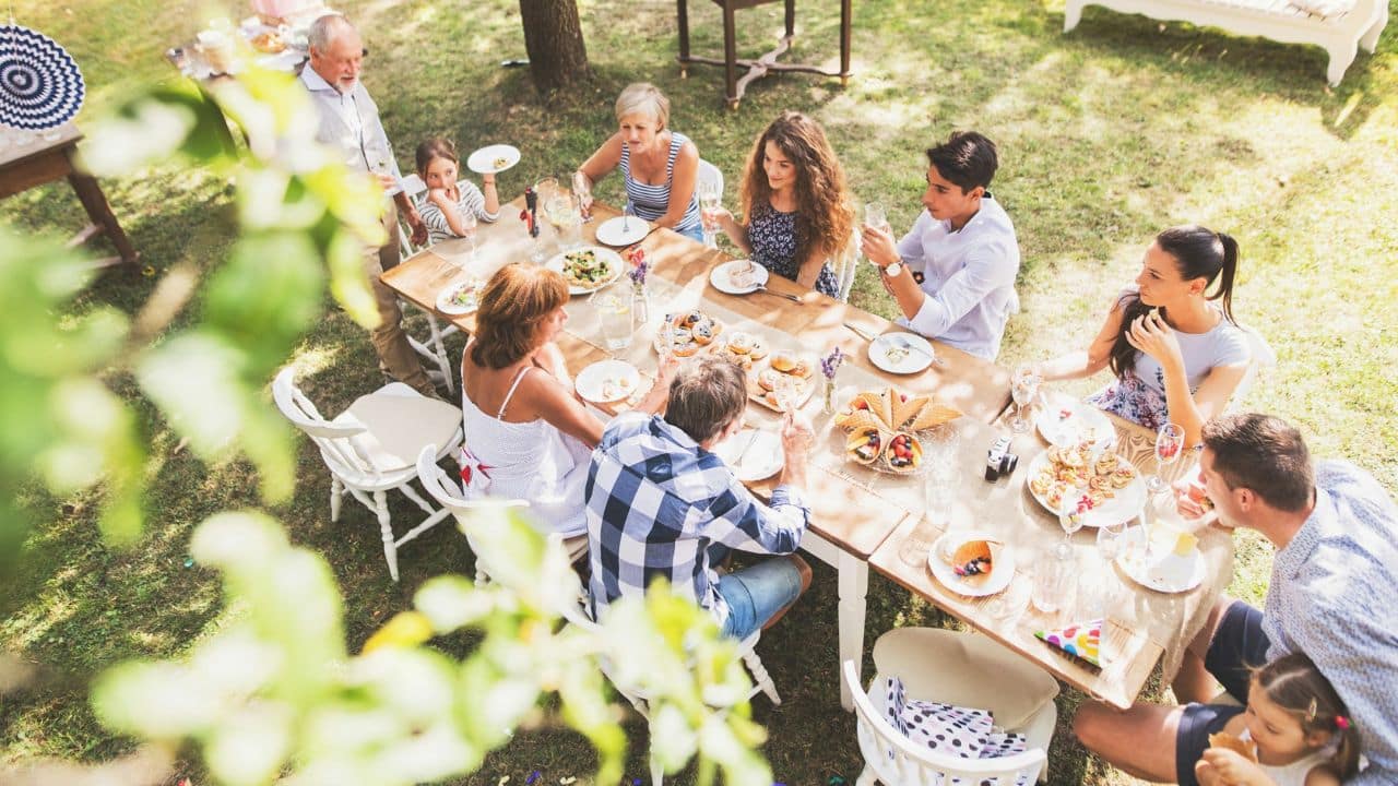 A group of family members of different ages is sitting around a table, enjoying a meal outdoors on a sunny day.