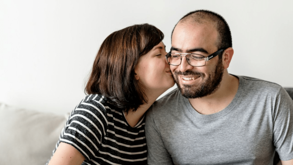 A young woman with brown hair kisses a smiling man on the cheek.