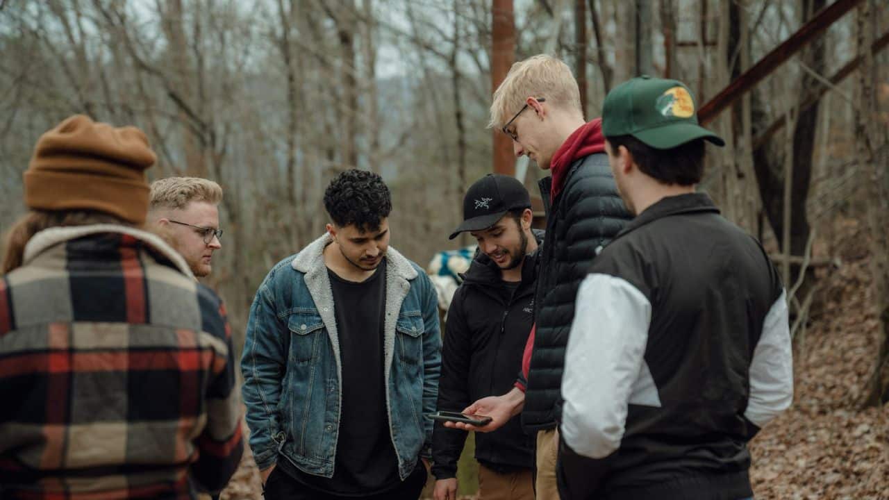 A group of five men stands in a wooded area, looking at a phone.