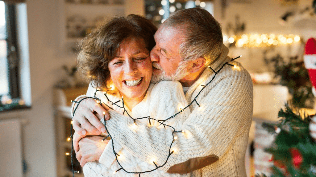 A laughing, older couple wrapped in holiday lights hugs affectionately.