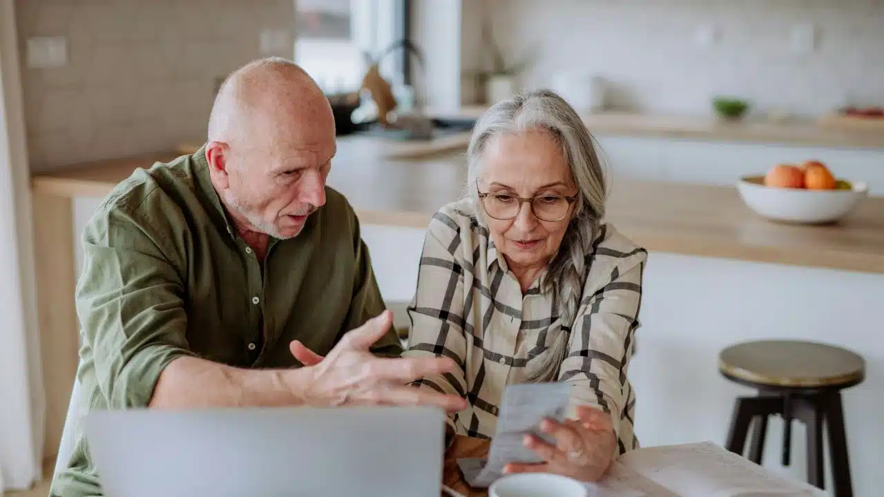 A mature couple sits at a kitchen table, looking concerned while reviewing a document.