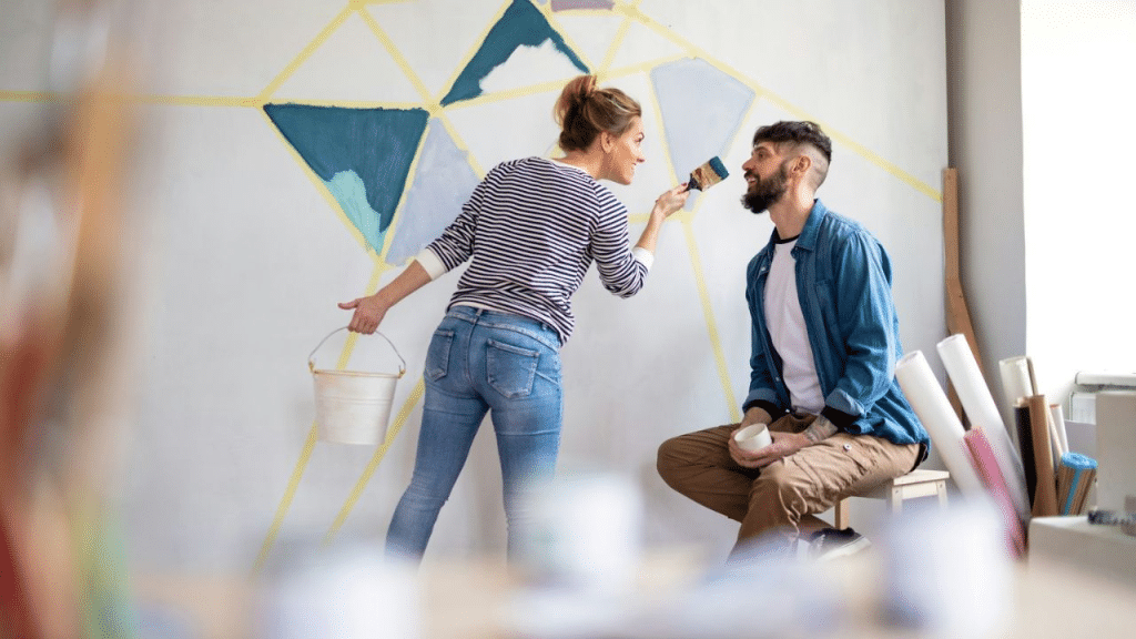 A smiling couple is playfully painting a wall and each other's faces.