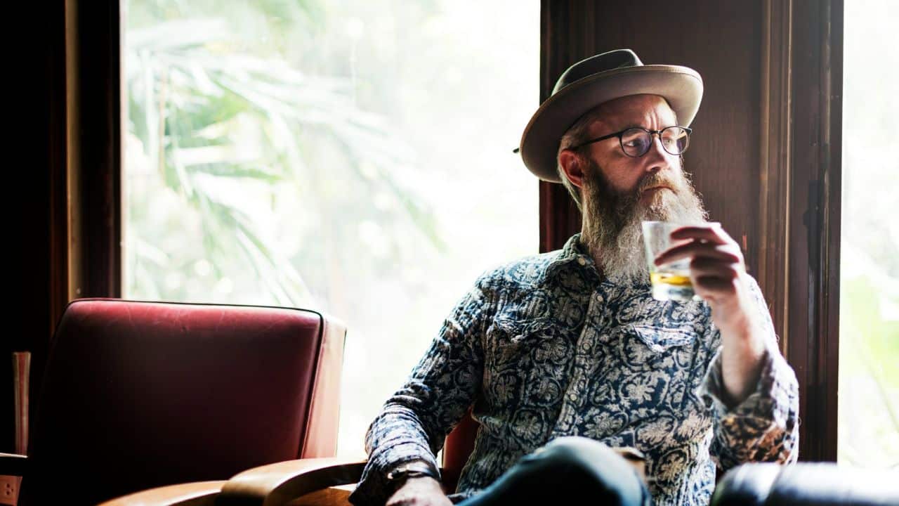 A middle-aged man with a beard and a hat sits in a chair by a window, holding a glass of amber liquid.