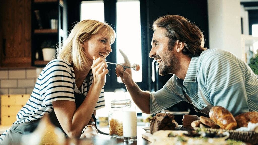 A joyful couple in a kitchen leans in close, laughing as the man points at the woman.