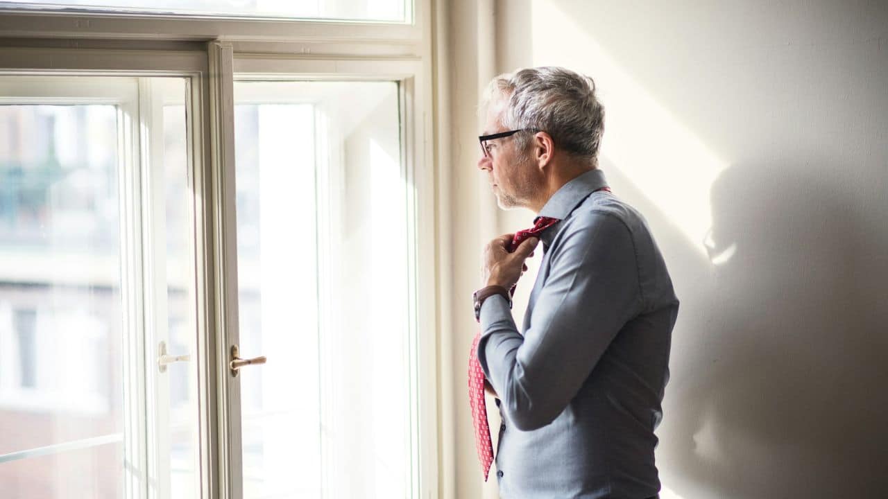 A gray-haired man in a button-down shirt stands thoughtfully by a window, adjusting his red tie.