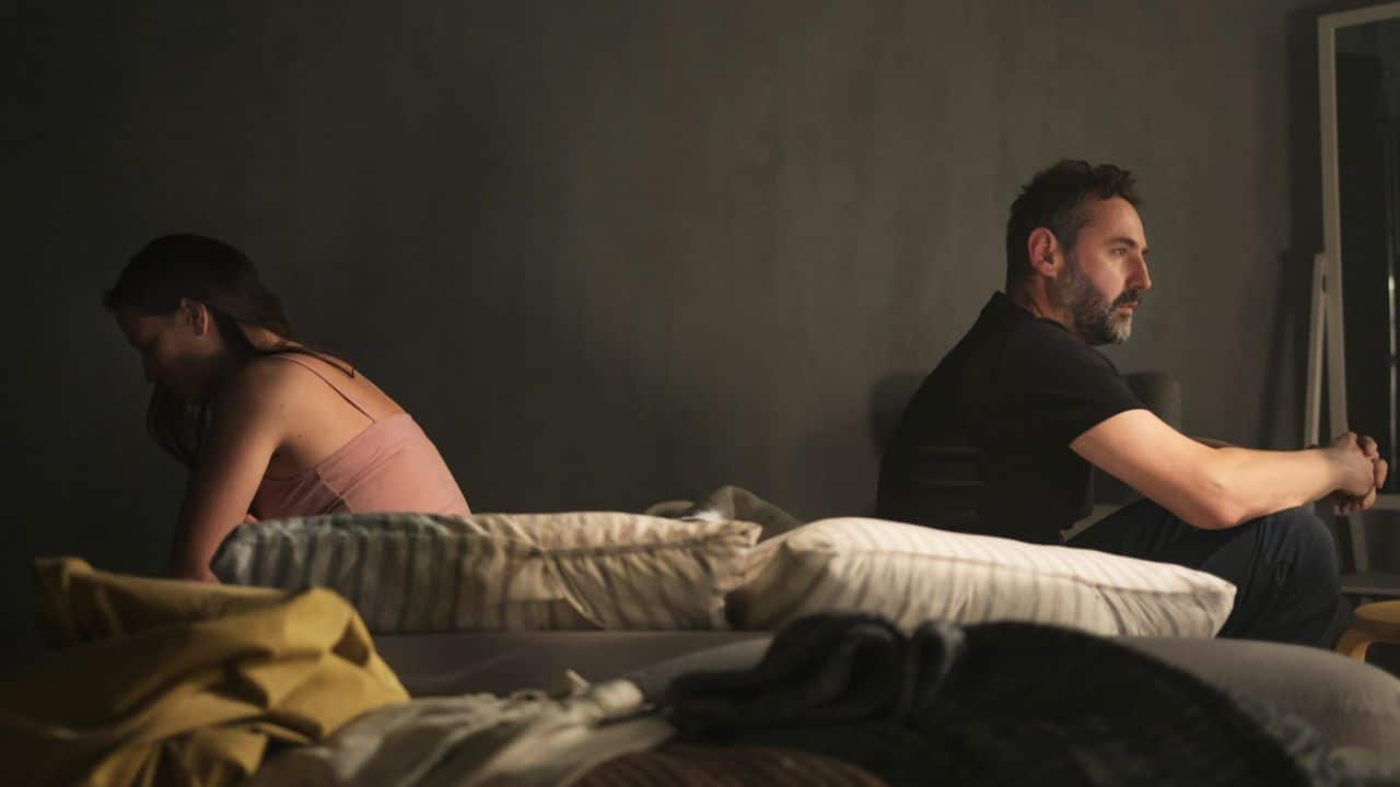 A sad-looking couple sits on a bed, back to back, with pillows between them.