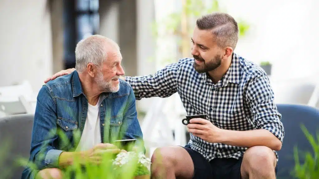 A millennial man with a checkered shirt is talking and smiling at a senior man in a denim jacket.