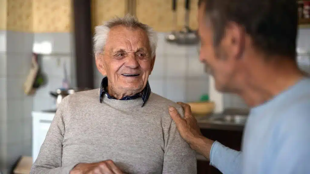 An older man in a tan sweater smiles warmly at a younger man with a hand on his shoulder.