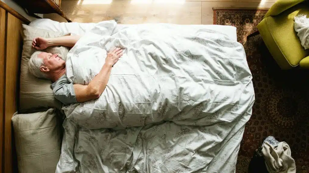 An overhead shot shows an older man asleep in his bed with a white comforter.