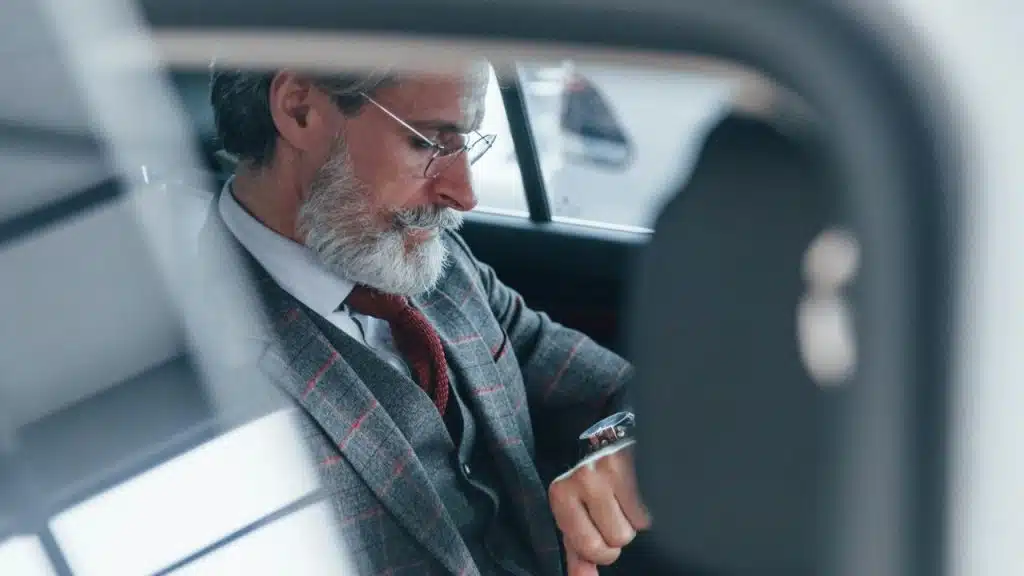 A senior man with a beard and glasses sits in the back of a car, looking at his watch.