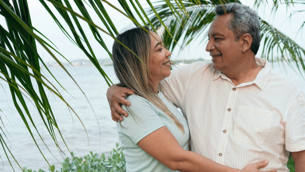 A mature couple stands outside near the water, with their arms wrapped around each other.