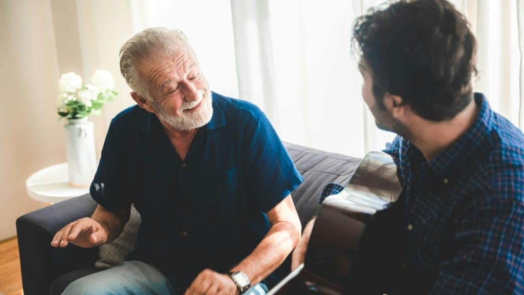 A smiling older man with a grey beard is seated on a sofa and listening to a younger man with a guitar.