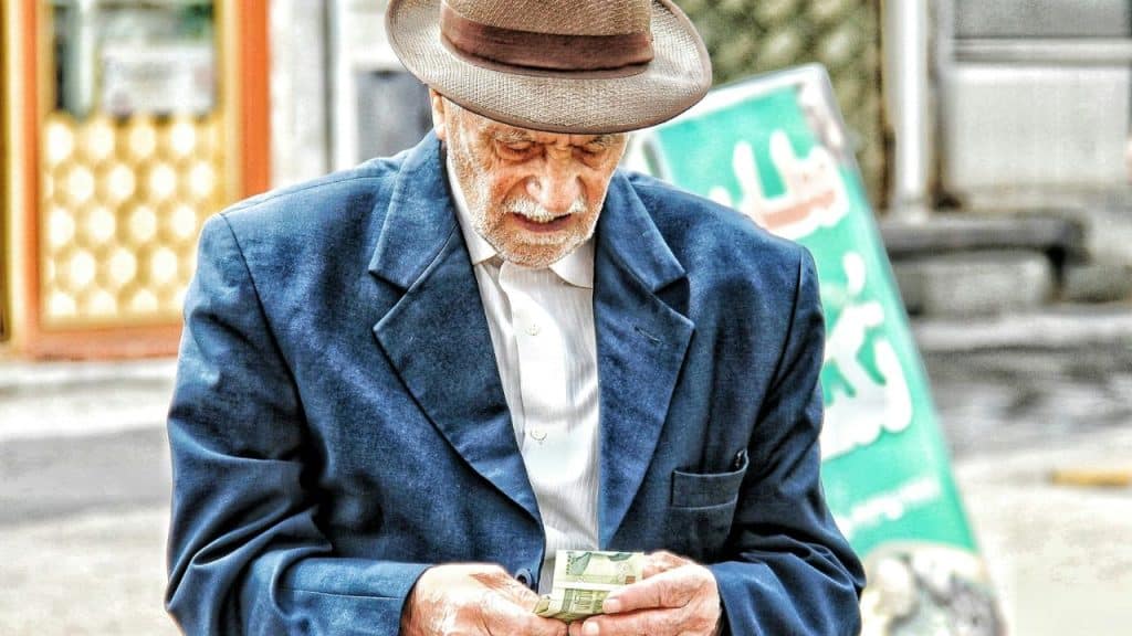 An older man in a fedora and blue jacket is looking down and counting money.