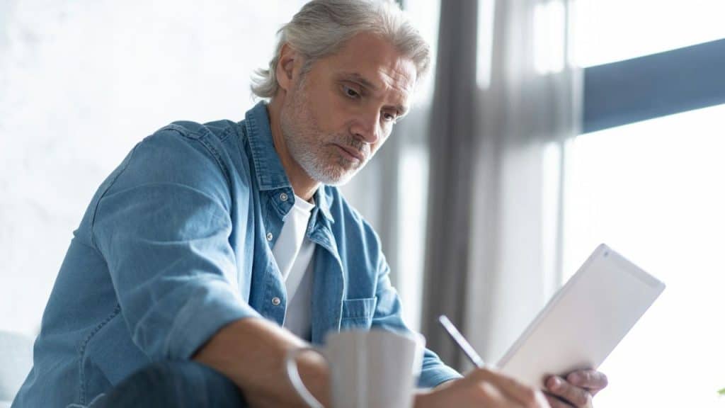 A senior man in a denim shirt is focused on writing something on a tablet with a stylus.