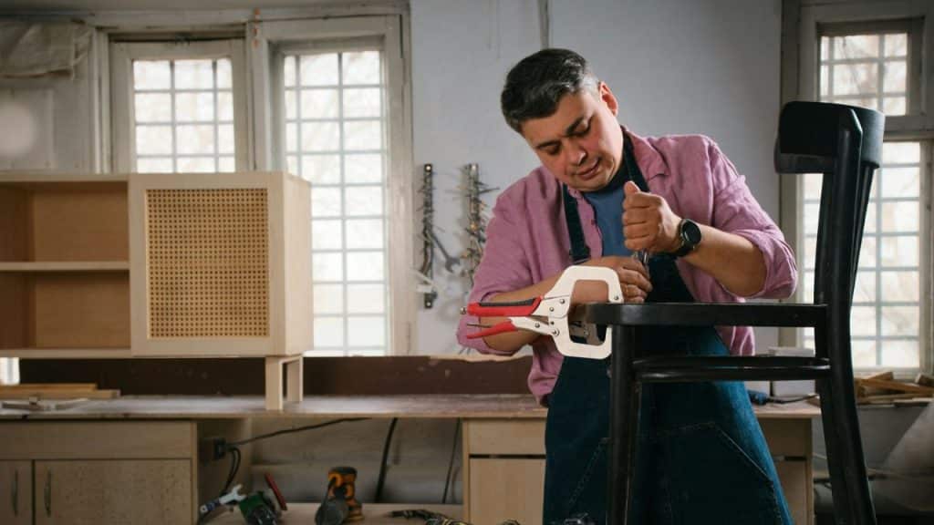 A craftsman wearing an apron is repairing a black chair with a clamp in a workshop.