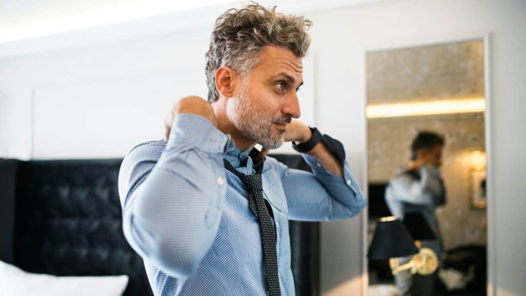 A handsome, middle-aged man with salt-and-pepper hair is adjusting his tie while looking in the mirror.