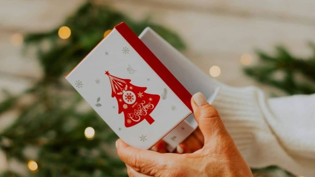 A hand is opening a small, white box with a red Christmas tree design on it.