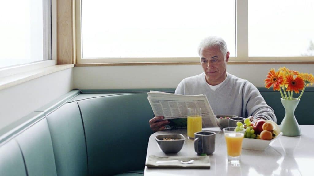A smiling older man is seated at a booth and reading a newspaper while having breakfast.