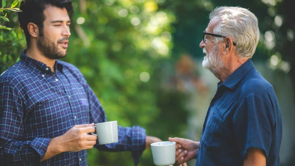 A senior man with glasses and a beard and a younger man with a beard are having a conversation and holding coffee mugs.