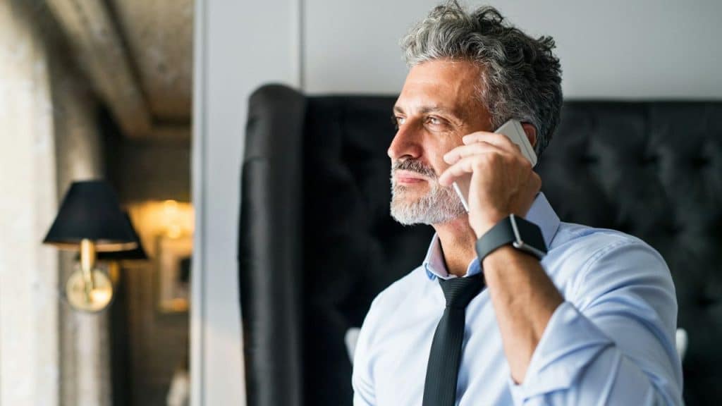 A man with grey hair and a beard is on a phone call while looking thoughtfully out a window.