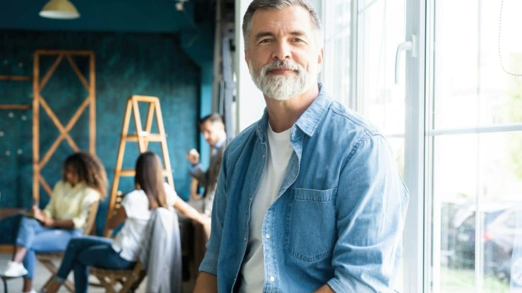 A confident, mature man with a beard and a denim shirt is smiling at the camera.