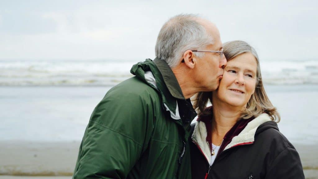 A middle-aged man in a green jacket is kissing a woman on the cheek at the beach.