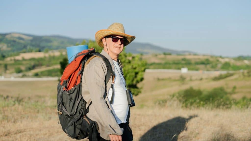 A Caucasian man in a hat and sunglasses with a large backpack is standing in a field.