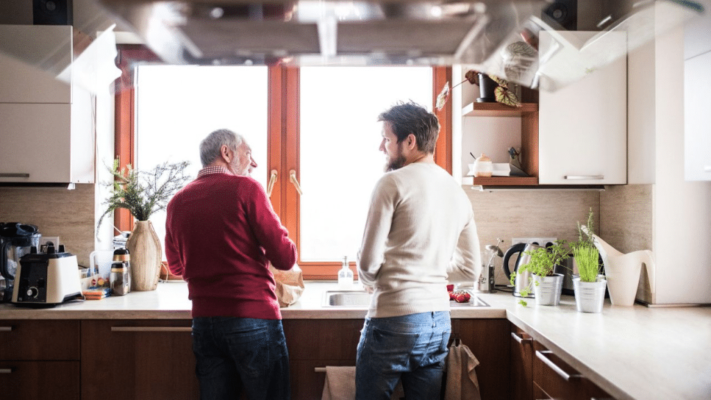 An older man and a younger man stand in a kitchen talking.
