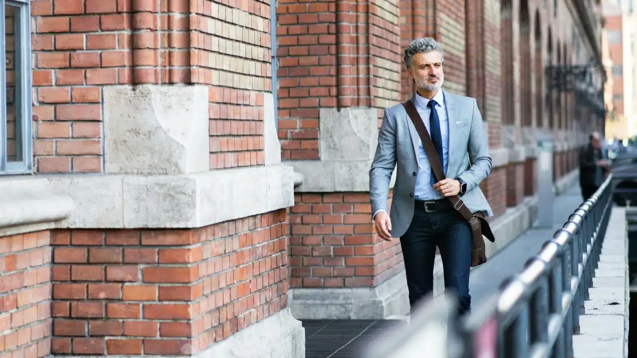 A man in a suit jacket and jeans with a messenger bag walks on a city sidewalk.