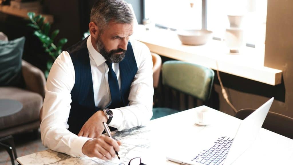 A focused businessman in a tie and vest is seated at a table, looking at a laptop and writing.