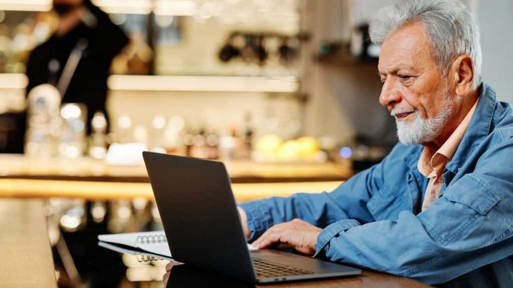A bearded older man in a blue jacket is seated in a cafe, looking at a laptop.