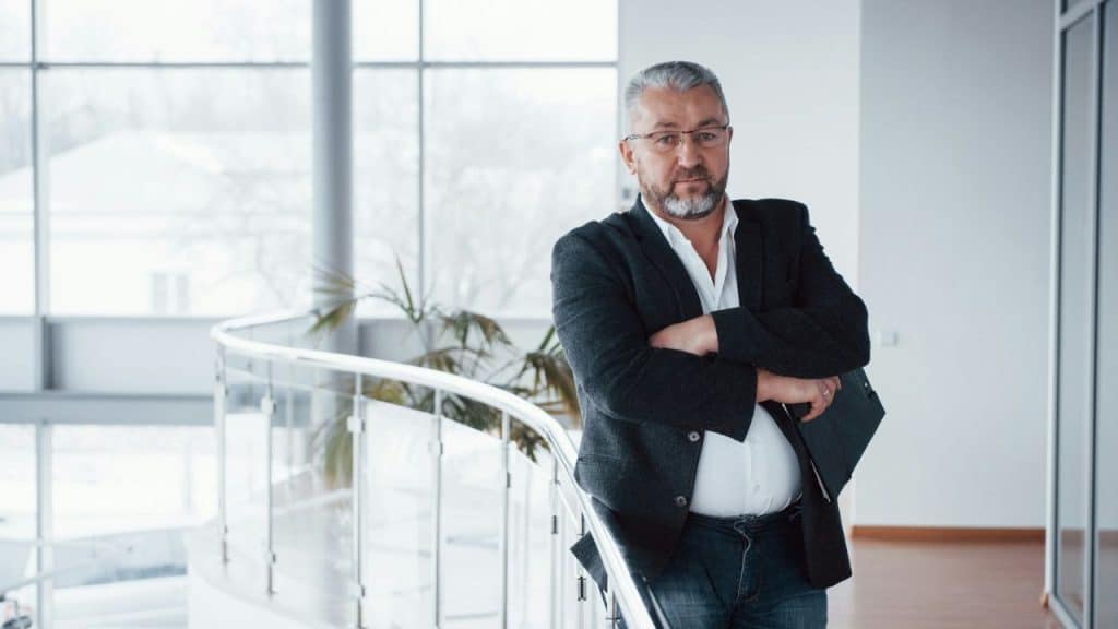 A senior businessman with glasses, a beard, and a gray jacket stands with his arms crossed.