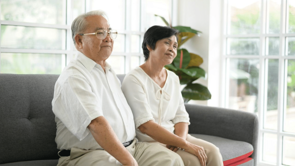 A middle-aged Asian couple sits together on a couch, looking out a window.