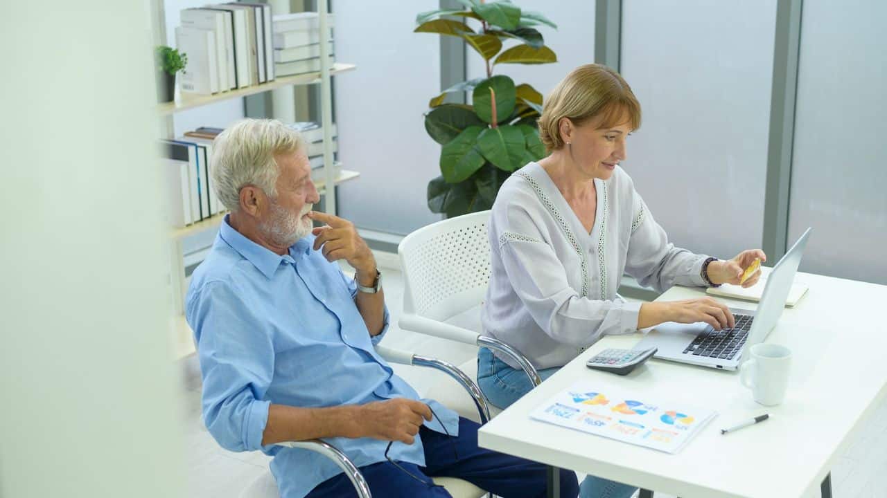 A middle-aged couple works together at a desk, with the woman using a laptop and a credit card while the man watches.