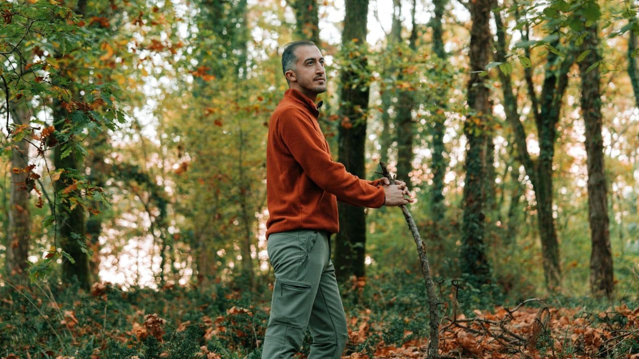A pensive man holding a walking stick stands in a forest with trees and fallen leaves around him.