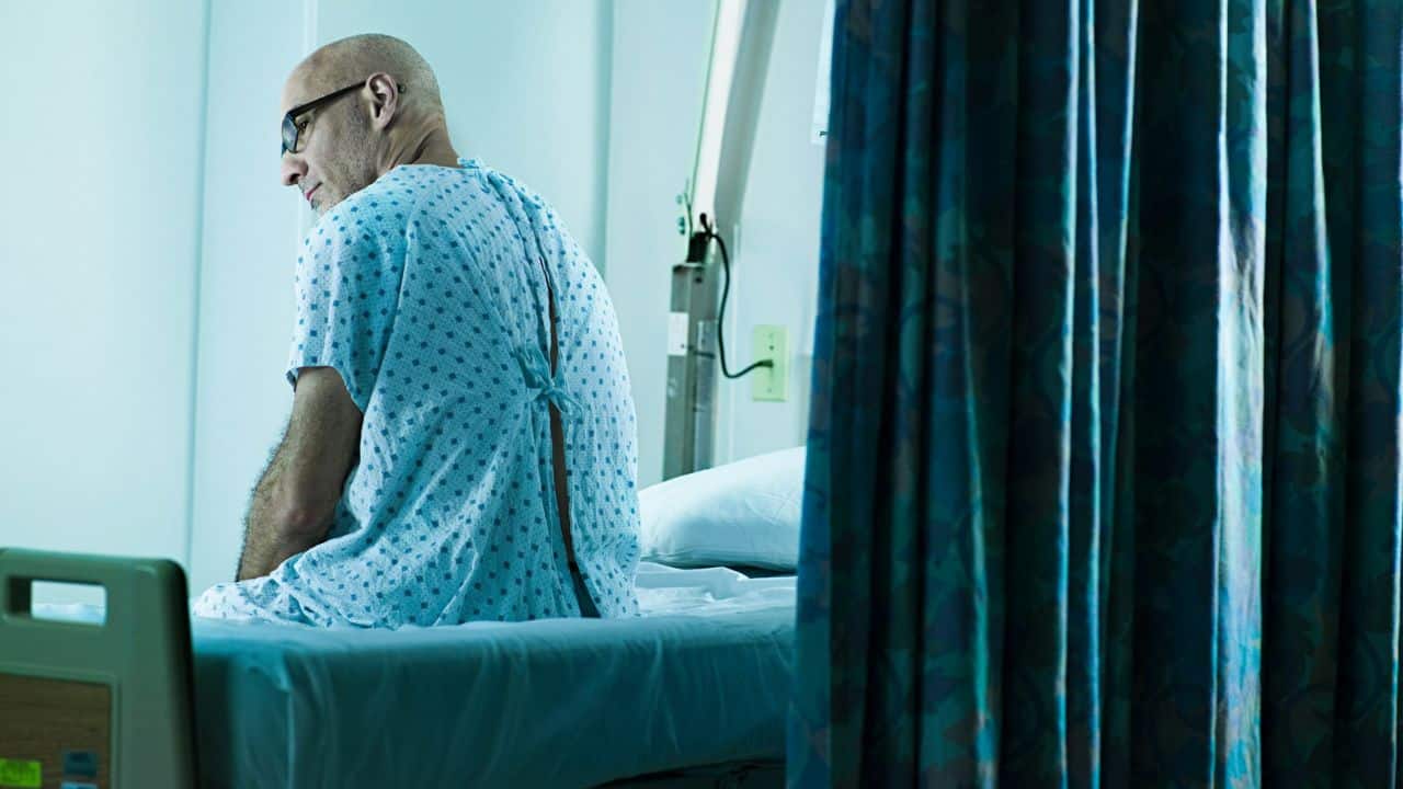A sad, bald man in a hospital gown sits on the edge of a bed, looking away.