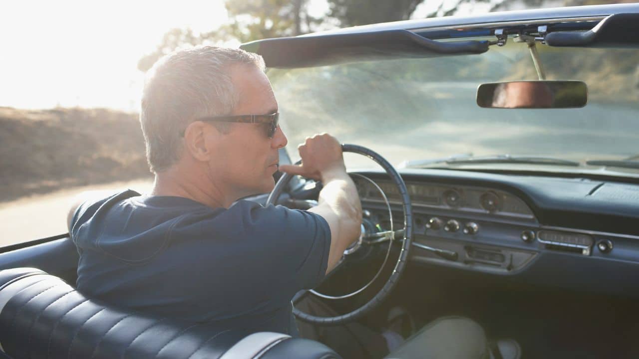 A gray-haired man in sunglasses drives a vintage convertible on a sunny day.