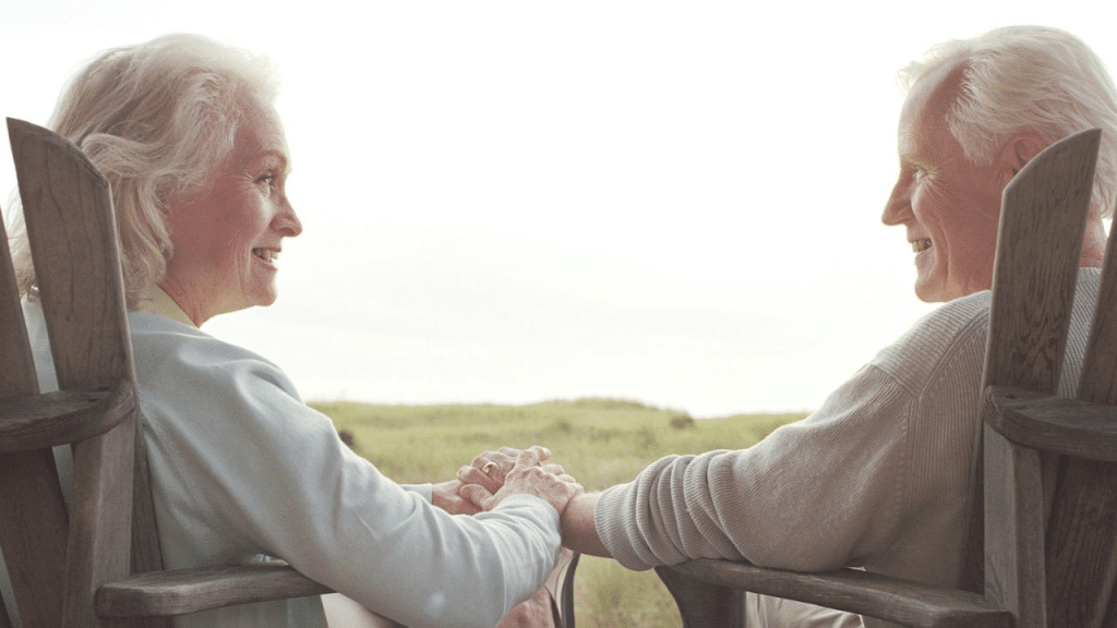 A cheerful, older couple holds hands while sitting in wooden chairs and smiling at each other.